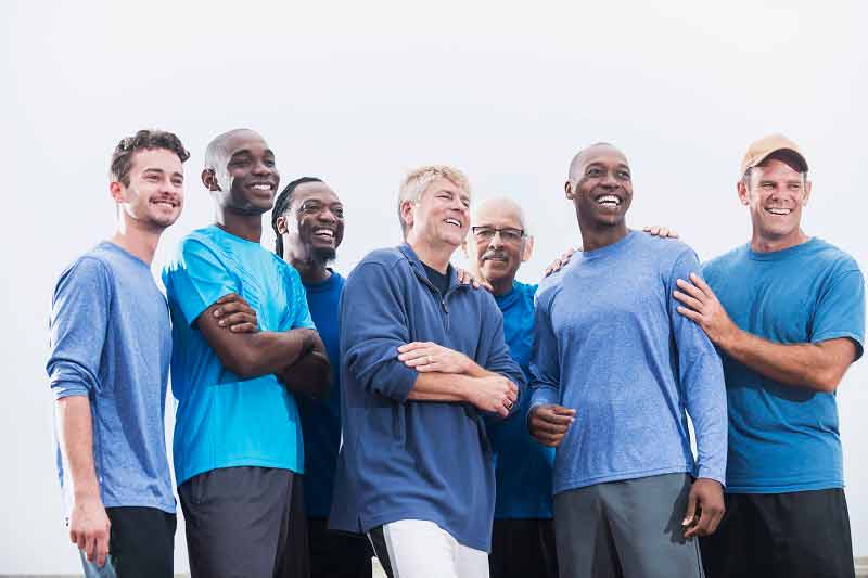 A group of 7 happy, healthy men of varying ages smiling and laughing outdoors.