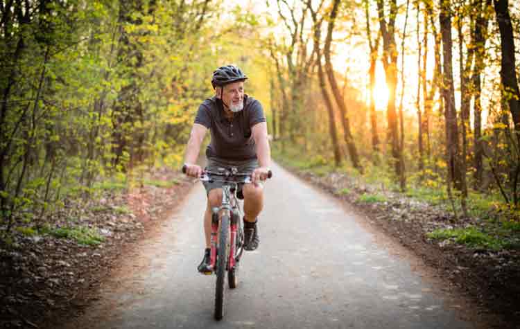 Older man riding his bicycle along a woodland road, with a low sun glowing warmly through the trees behind him.