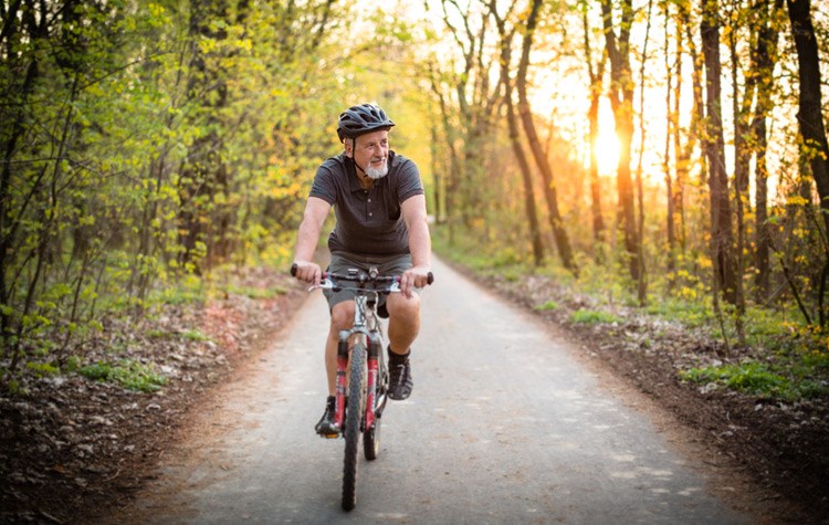 Older man riding his bicycle along a woodland road, with a low sun glowing warmly through the trees behind him.