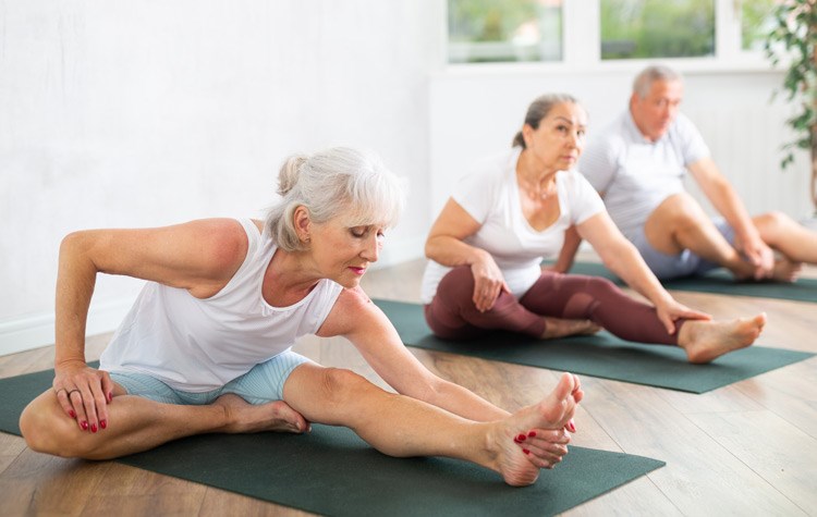 Senior visitor of yoga class and young female couch engaged Pilates in sports studio hall. Elderly woman stretches and prepares