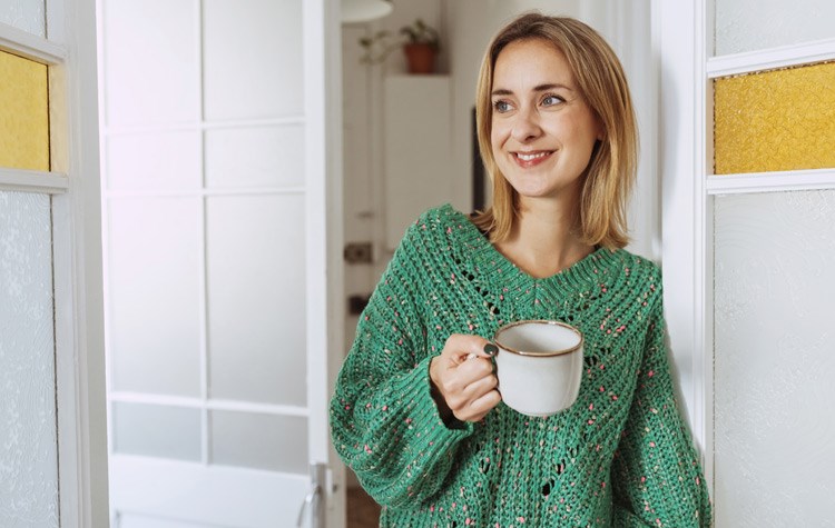 Young beautiful woman with cup of coffee, wears a green sweater, looks away at home.
