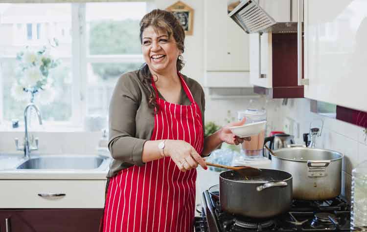 Mature woman is talking and laughing with someone out of the frame while she makes dinner at the cooker.