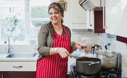 Mature woman is talking and laughing with someone out of the frame while she makes dinner at the cooker.