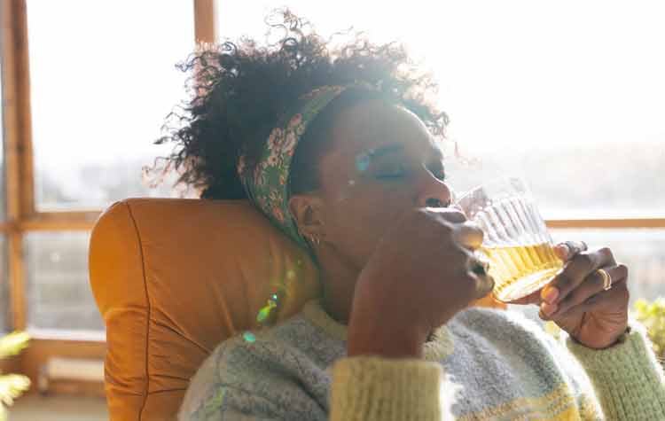Woman relaxing laying in a chair drinking a cup of tea at her home. She has her eyes closed and the sun is shining through the windows behind her.