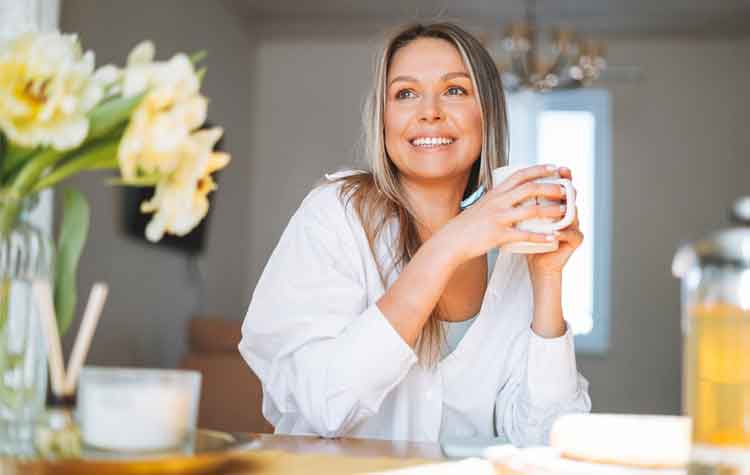 Young, happy woman with blonde long hair in white shirt drinking tea with bouquet of yellow flowers in vase 